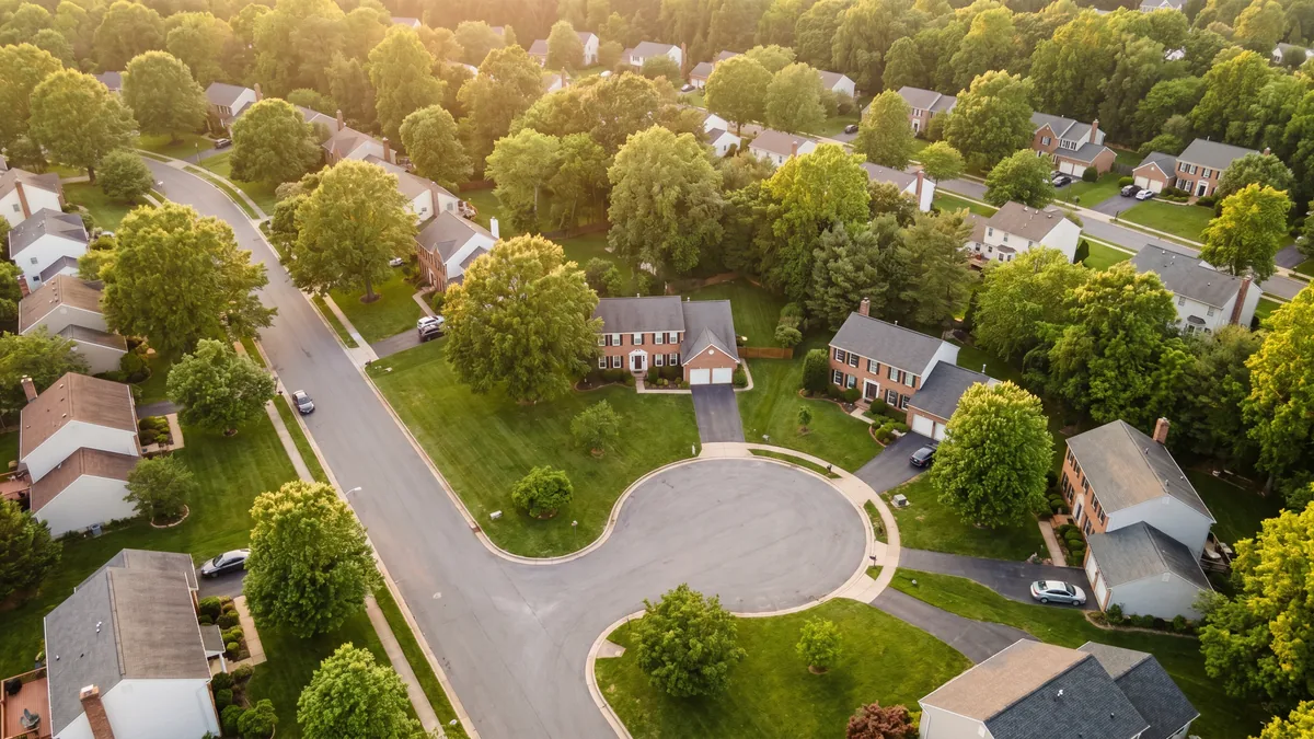 Aerial view of an established Maryland suburban neighborhood with detached single-family homes, a cul-de-sac, and a dense mature hardwood canopy at golden hour